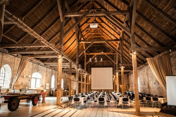 Rustik lade med højt til loftet, synlige bjælker og opsat til konference med borde og projektor – unikke eventfaciliteter på Sonnerupgaard Gods