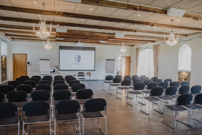 Stort konferencelokale med rækker af stole, projektor og træbjælker i loftet – ideelt til kurser og foredrag på Sonnerupgaard Gods