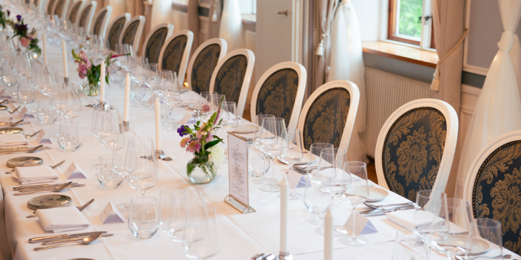 Langbord dækket op til sølvbryllup med hvide duge, levende lys, blomster, vinglas og elegante stole i festlokale på Sonnerupgaard Gods