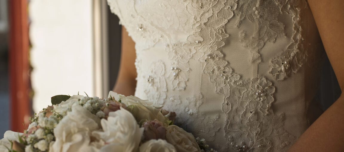 Detaljebillede af brudekjole med blonder og brudebuket med hvide og lyserøde blomster